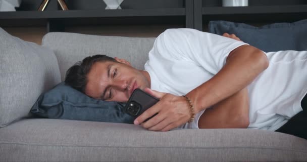 抑圧された男はソファーの上に横たわり 携帯電話のフィードをスクロールします うつ病や疲労について 携帯電話の画面と思考で主演する失望したアパテチック男性 — ストック動画