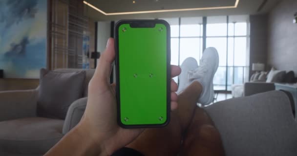 年轻男子躺在沙发上 家里有垂直模式的绿色屏幕智能手机 男性在客厅手持绿色屏幕智能手机 浏览互联网 观看视频内容 — 图库视频影像