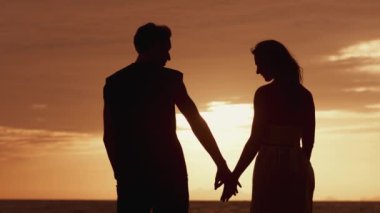 Gün batımında birbirini seven bir çiftin silueti. Aşık bir adam bir kadını elinden tutar ve denizdeki altın gün batımını seyreder. Gün batımında aşıkların romantik sahnesi. Yaz romantik tatilleri.