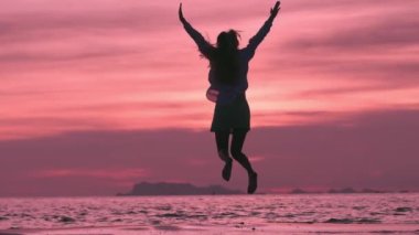 Pembe gün batımında sahilde zıplayan mutlu kadın turist. Yavaş çekimde sevinçten zıplayan genç bir kadının silueti. Mutlu turist kadın denizde yaz tatilinin tadını çıkarıyor. Mutluluk ve başarı kavramı