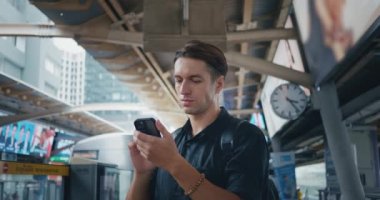 Genç adam metro istasyonunda yaklaşan treni beklerken akıllı telefonu kullanır. Toplu taşıma durağındaki 30 'lu yılların yolcusu modern şehirde tramvay bekliyor. Teknolojik ulaşım.