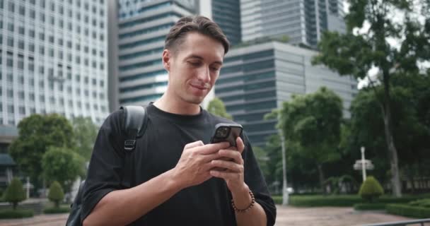 スマートフォンを使ってバックパックで笑顔の男学生 スマートフォンを閲覧する男性学生観光客は 建物の背景に大きな都市でアプリや地図を見ます — ストック動画