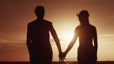 Arkadan bakıldığında, bir erkek bir kadının elini turuncu bir günbatımının arka planına koyar. Gün batımında balayında yolculuk eden sevgi dolu bir çiftin silueti. Bir erkeğin bir kadını sevmesi kavramı.