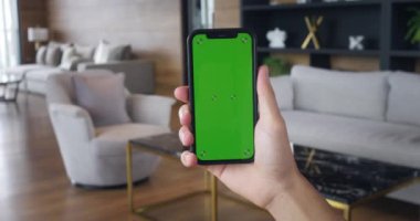 Genç adam oturma odasında dikey yeşil ekranlı akıllı telefonu tutuyor. Evde yeşil ekran krom tuşlu akıllı telefon kullanan erkek modeli. İnternet taraması, Video İçeriği İzleme, Bloglar.