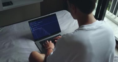 Dijital Çalışma Alanı. Dizüstü bilgisayarda Adam Daktilo Kodu, Yatak ve Arka Görünüm 'de oturuyor. IT Freelancer yatağının konforundan bilgisayarına kod yazıyor..