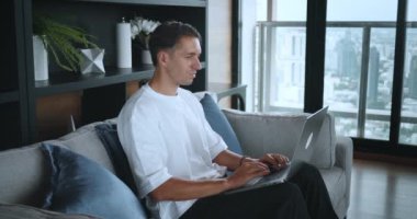Genç adam kanepede oturuyor, evden bilgisayarla çalışıyor ya da ders çalışıyor. Erkek öğrenci dizüstü bilgisayar kullanarak öğreniyor, internetten öğreniyor. Uzaktan eğitim kavramı, serbest çalışma, ev kavramından uzak çalışma.