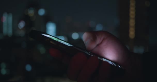 閉じる人は都市の光の背景で夜間に携帯電話を使用します 男性の手のシネマティックフィルムの外観は 携帯電話を使用し 夜に彼の携帯電話上のアプリ上でニュースフィードをタップしてスクロールします — ストック動画