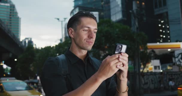微笑的恋爱中的男人站在城市的夜晚街上 一边用手机给女朋友留言 市民可透过流动电话交换讯息 在市区灯光下进行互联网通讯 — 图库视频影像