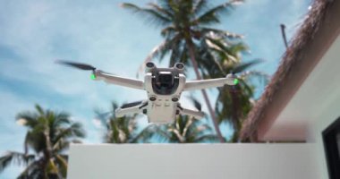 Mavi yaz gökyüzünün arka planında havada süzülen modern insansız hava aracı, tropik tatil beldesinde palmiyeler. Yaz tatilinde seyahat blogcusu içeriğini kaydetmek için modern drone helikopteri.