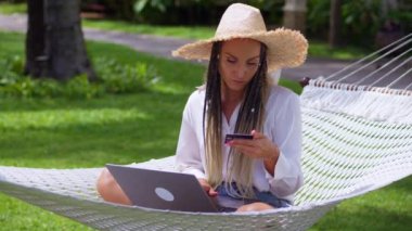 Çevrimiçi bankacılık. Hasır şapkalı bir kadın kredi kartıyla internet üzerinden alışveriş yaparken yaz seyahatinde hamakta dinleniyor. Yaz tatilleri sırasında kredi kartlarıyla online alışverişler