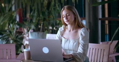 Kafeden laptopta çalışan genç bir kadın. Kafede çalışan sarışın bir kadın. Bilgisayarda mesaj yazıyor. Kafeteryada çalışan beyaz bir kadın, rahat bir yerde oturuyor ve kahve içiyor..