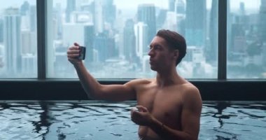 30 'lu yaşlardaki bir adam tatilde otelin havuzunda cep telefonundan videoyu arıyor. Gömleksiz erkek kapalı gökdelen havuzunda görüntülü konuşma yapar. Gülümseyen turist lüks otel havuzunda video görüşmesi yapıyor..