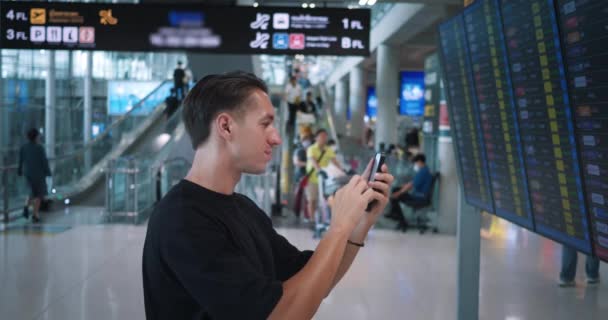 手持智能手机的人类旅行者在查看到达和离开信息时 会显示对其航班的搜索 白人旅客在飞行信息板 检查航班 旅行概念 — 图库视频影像