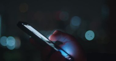 Kapanan kişi, gece vakti şehrin aydınlık arka planında akıllı telefon kullanır. Erkek elinin sinematik görüntüsü, akıllı telefon kullanımı, gece telefonundaki uygulamayı dinlemek ve kaydırmak..