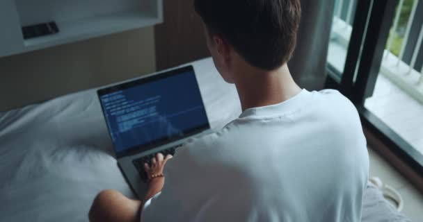自宅でベッドに座っている間 ラップトップ上でソフトウェアコードを作成する男のリアビュー フリーランスの男は スクリーンラップトップ上のコードとプログラミングをタイプします ハッカーは自宅からオンラインで仕事をしている ラップトップ上のコードを作成する男 — ストック動画