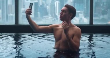 İş adamı tatilde otelin havuzunda telefonla videoyu arıyor. Kapalı gökdelen havuzunda tişörtsüz iş adamı görüntüsü. Gülümseyen adam lüks otel havuzunda iş videosu çekiyor..
