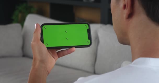 リビングルームで横の緑色のスクリーン電話でスマートフォンを握る肩のショットマンの上 自宅で緑色の画面をモックアップしてスマートフォンを使用する男性の手 インターネット閲覧 ビデオを見る — ストック動画