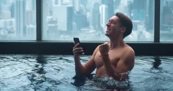 快乐的30多岁的男人享受着成功的喜悦 一边在智能手机里读好消息 一边在城市背景下的摩天大楼屋顶上的豪华游泳池里休息 流动彩票胜出概念 — 图库视频影像