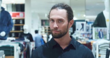 Kıyafet mağazasında dikilip gülümseyerek kameraya bakan yakışıklı bir adamın göğüs portresi. Giyim departmanının modaya uygun kıyafetlerine karşı sakallı şık bir adam.