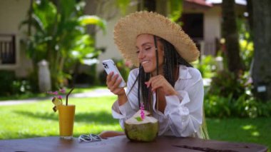 Tatil köyündeki kadın telefonda komik videolar izliyor ve meyve suyu içmek mutluluk saçıyor. Bir kadının yaşadığı samimi mutluluğu hissedebilir. Dertsiz ve mutlu tatil kavramını tropik tatil beldesinde.