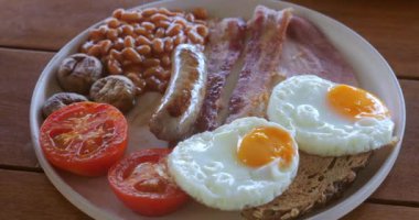 Tabakta İngiliz kahvaltısı, yakın çekim. Yağda yumurta, sosis, pastırma, domates, fasulye, ekmek kızartması, ahşap masada beyaz tabakta. Günaydın Avrupa kahvaltısı.