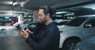Sosyal ağlarda gülümseyen bir adam telefonda, otoparktaki mutlu adam telefonundaki arkadaşlarının ona gönderdiği görüntülere hevesle bakıyor. Pozitif telefon iletişimi kavramı.