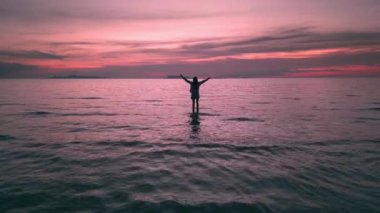 Gün batımında cennet adasındaki tropikal sahilde genç ve mutlu bir kadın. Gün batımında siluet kadın kollarını havaya kaldırıyor ve tropik cennet adasında gün batımını selamlıyor.