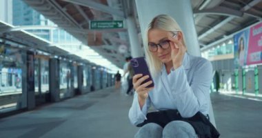 Metro istasyonunda bekleyen akıllı telefon kullanan bir kadın. Şehir dışında metro istasyonunda bekleyen bir kadın. Ev ödevi üniversite çalışmalarına giden yol haritasını kontrol ediyor. Mutlu kadın oturmuş mesaj yazıyor..
