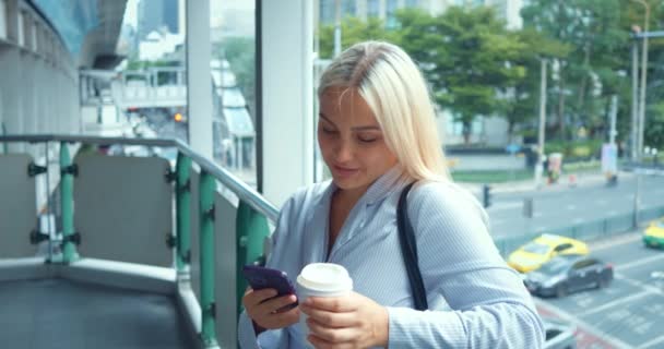 市内を歩いている女性が 電話を使って ポートレート ミレニアル世代の女性 コーヒーカップと忙しい道路の背景に携帯電話 リラックスしてブリッジに立ち 微笑むタイピングメッセージ — ストック動画