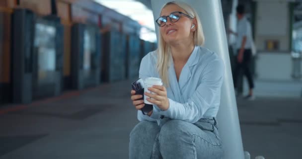 地下鉄で音楽を聴いている女性 若い幸せは 地下鉄駅でコーヒーカップスマートフォンのスカウトを保持するワイヤレスヘッドフォンのイヤホンで音楽に揺れる幸せなミレニアル女性を満足させました — ストック動画