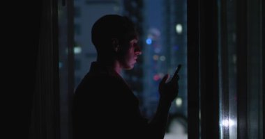Gecenin geç saatlerinde elinde telefonla gökdelenin penceresinde duran bir adam. Millennial uzun bir iş gününden sonra telefonda mesajları okur. Şehir ışıkları ve telefon ışığında yakışıklı bir adamın sinematik görüntüleri.