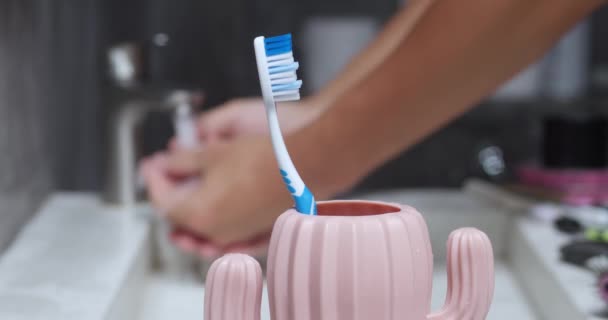 人洗手 牙刷在前景 牙刷杯的形状为粉色仙人掌 蓝白相间的牙刷 现代水槽中的人洗手 背景模糊 有选择的重点 — 图库视频影像