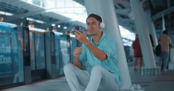 イヤホンを着ている若者は 駅のプラットフォームで地下鉄を待っていて 電話で話している アクティブな男は電話でオンライン交渉を行います 高速都市生活の現実における現代人の概念 — ストック動画