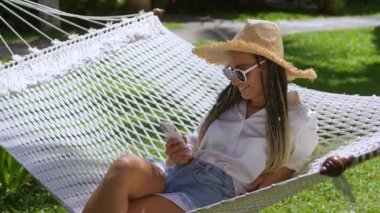 Hasır şapkalı ve güneş gözlüklü kadın gezgin yaz otelindeki hamakta uzanıyor ve elinde akıllı bir telefon tutuyor. Kadın gezgin hamakta dinlenmeyi ve yaz serinliğinde akıllı telefon uygulamasını kullanmayı sever..