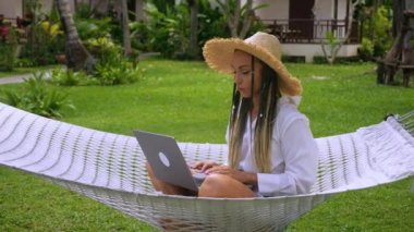 Hasır şapkalı iş kadını gezgin yaz tatilinde hamakta çalışmak için dizüstü bilgisayar kullanıyor. Hasır şapkalı kadın hamakta dinlenirken dizüstü bilgisayarda çalışıyor yaz tatilinde..