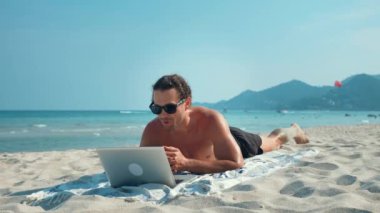 Güneş gözlüklü plaj adamı bilgisayarla iletişim kuruyor. Video çağrısının gücü, iş ve eğlence esnekliğini bir araya getiriyor. Tranquil plaj ayarı, uzak çalışma video çağrısının rahatlığı.