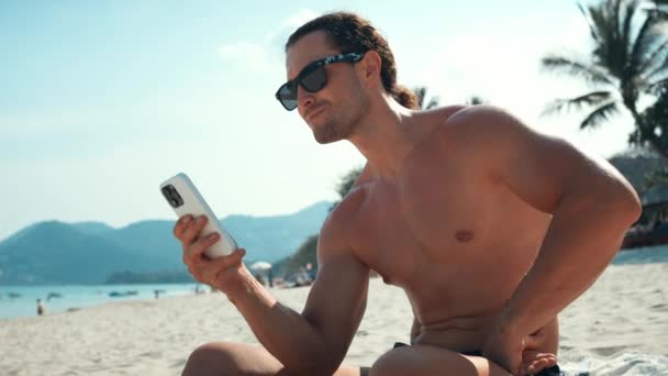 Uomo Senza Maglietta Occhiali Sole Siede Sulla Spiaggia Utilizzando Telefono — Video Stock
