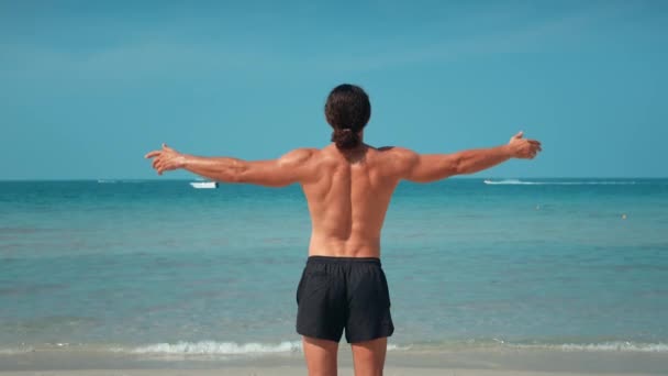 バックグラウンドの筋肉質男性ポーズの海は ビーチを立っています ビーチリラクゼーションアスレチックな体格と決意チャンピオンポーズのブレンドを象徴するチャンピオンのポーズを反映した上げられた腕 — ストック動画