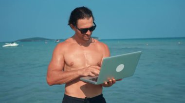Sahilde ayakta duran güneş gözlüklü bir adam okyanus esintisi ile dizüstü bilgisayarlı göçebe üzerinde çalışır ve çalışmanın ve boş zamanların dijital göçebe olduğunu sembolize eder. Dijital göçebe modern teknoloji çalışması