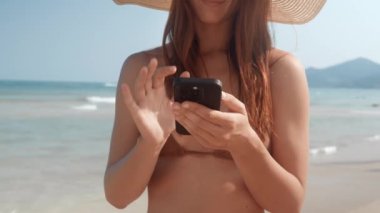 Bikini giyen ve plajda şapka takan yakın plan kadın sosyal ağların cep telefonunda geziniyor. Kadın telefon ekranına bakıyor, haber akışında kayıyor. Telefon, turist plajın tadını çıkarırken