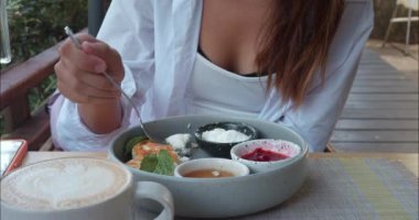 Genç bir kadın açık verandada rahat bir yazlık kafede kahvaltı yapıyor. Kadınlar lezzetli peynirli kekleri ve kafeden büyük bardak kapuçinoyu sever. Yaz kafesi atmosferi ve lezzetli kahvaltı zevki.