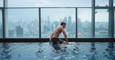 Milenyum erkek turist, lüks bir şehirde lüks bir otelde, çatı üstü yüzme havuzunun kenarında dinleniyor. Genç adam şehrin arka planındaki gökdelenin çatısında lüks bir yüzme havuzunda eğleniyor..