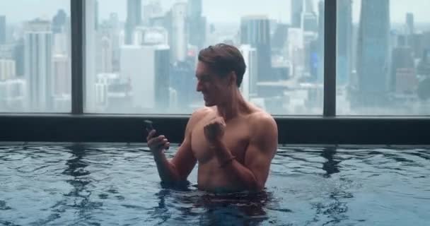 在大城市 富人使用智能手机 在摩天大楼的顶楼游泳池里放松一下 具有城市背景的豪华酒店顶层游泳池 手持智能手机的千年男性游客 — 图库视频影像