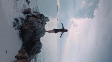 Okyanus kıyısındaki bir kayanın üzerinde duran dikey videolu kadın, kolları açık deniz rüzgarı. Okyanustan esen güçlü rüzgar saçlarını dalgalandırır ve elbiseni dalgalandırır. Kadınlar el değmemiş ve güçlü okyanus elementlerini sever.