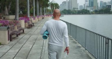 Üst düzey bir adam yoga paspasıyla parka doğru yürüyor ve mükemmel bir yer arıyor. Yoga seansı için şehir parkında yürüyen adam işe gitmeden önce sabah antrenmanı yapıyor..