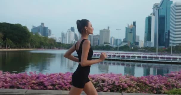 在城市公园的摩天大楼中 女运动员优雅地跑着小径 平静地跑着湖水 快乐地跑着健康的生活方式 夏日的微风 精力充沛的女人跑步 激励着她的健身之旅 — 图库视频影像