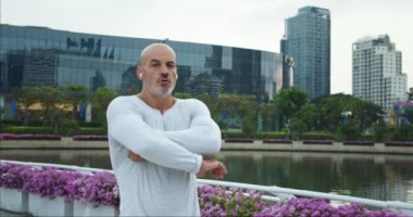 Şehir parkında yakışıklı, beyaz, yaşlı bir adam kollarını kavuşturmuş kameraya bakıyor kendinden emin bakışlar. Olgun insan kendine güveni olan iç güç soğukkanlılığı dingin atmosfer parkında yankılanıyor
