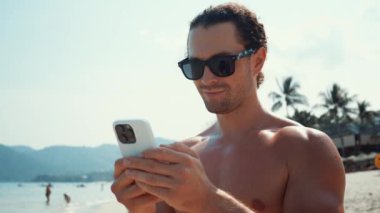 Millennial, tropik adanın sıcak güneşinin altında sahilde duruyor ve komik videoların telefon görüşmelerini izliyor. Adam komik içerikli telefon ekranına bakıyor. Telefon, doğal cennetteki dijital dünyaya bağlanıyor.