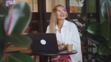 Orta yaşlı sarışın bir iş kadını güzel bir kafede oturuyor ve verimli bitkiler dizüstü bilgisayarla çalışıyor. Orta yaşlı, beyaz, sarışın, iş kadını. Göz kamaştırıcı bir kafede oturuyor. Yeşillik de bilgisayarla çalışıyor.