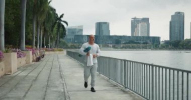 Atletik vücutlu bir adam sabahları yoga minderi ve havluyla şehir seti boyunca spor yapmaya gider. Açık hava aktiviteleri için uygun bir zaman. Eğitimden önce sabah yürüyüşü insanın sabah ritüelidir.